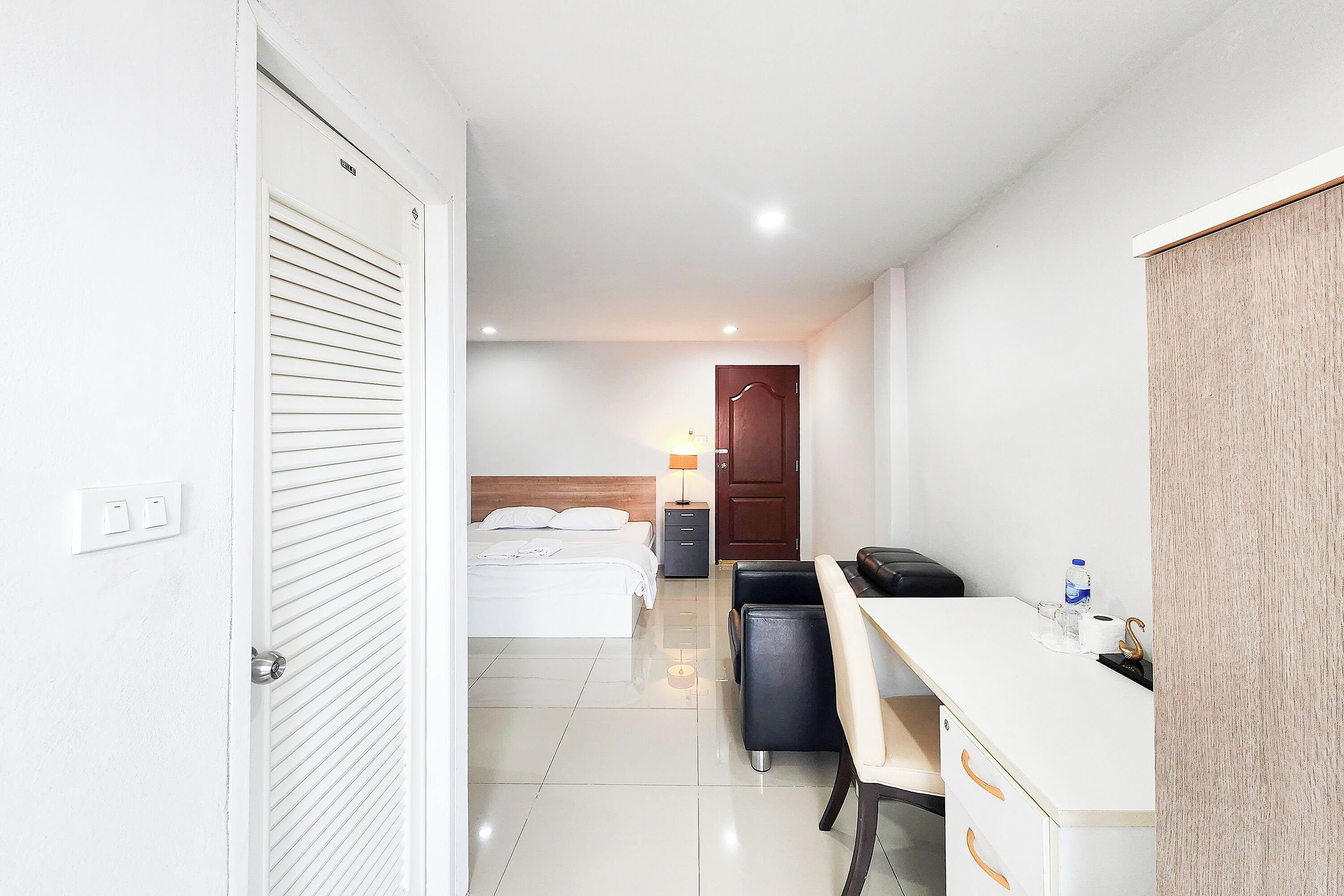 A hotel room with a bed, desk, chair, and a small table with a lamp and a bottle.