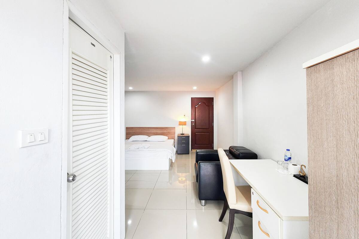 A hotel room with a bed, desk, chair, and a small table with a lamp and a bottle.