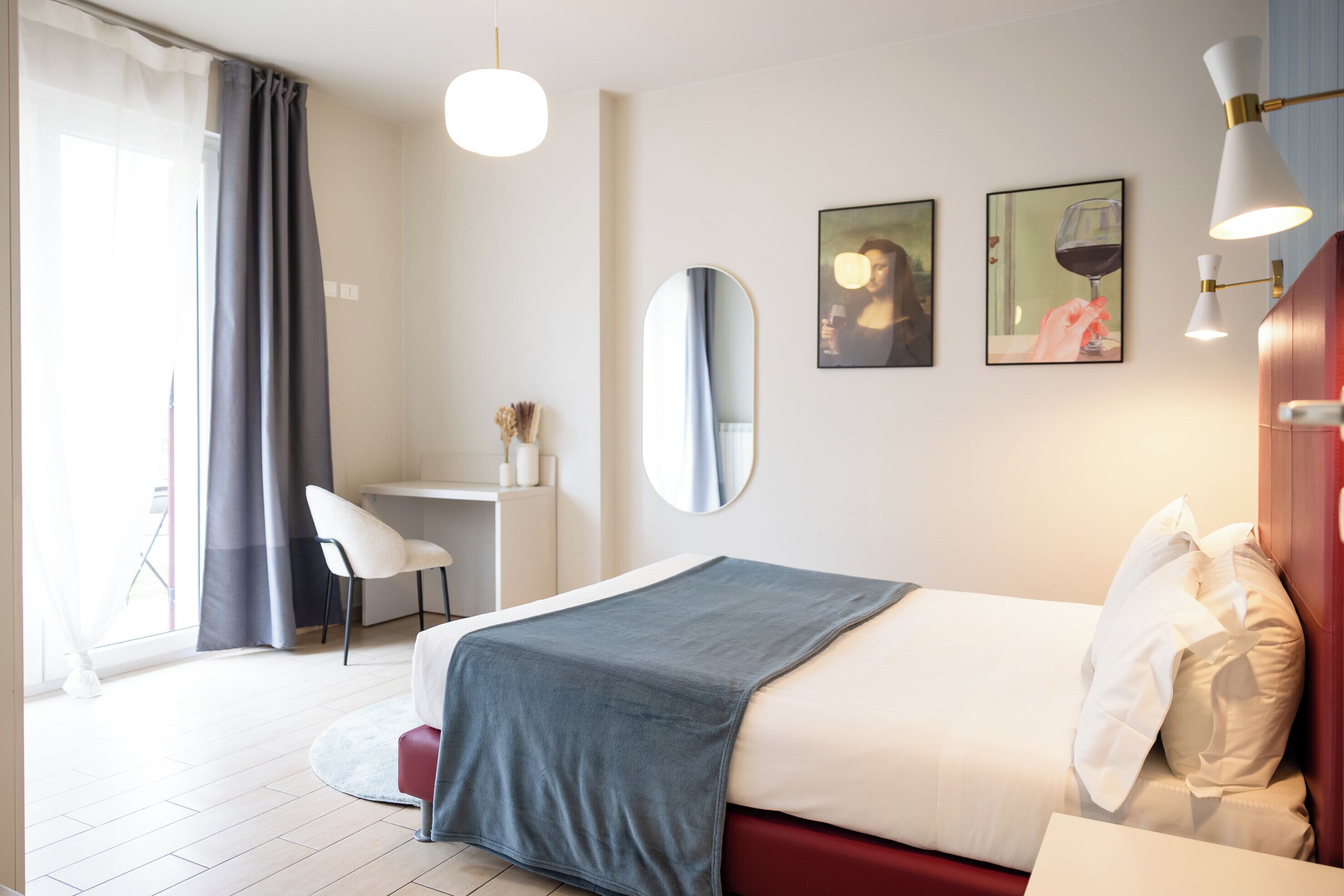 A hotel room with a bed, a desk, a chair, a mirror, and artwork on the wall.