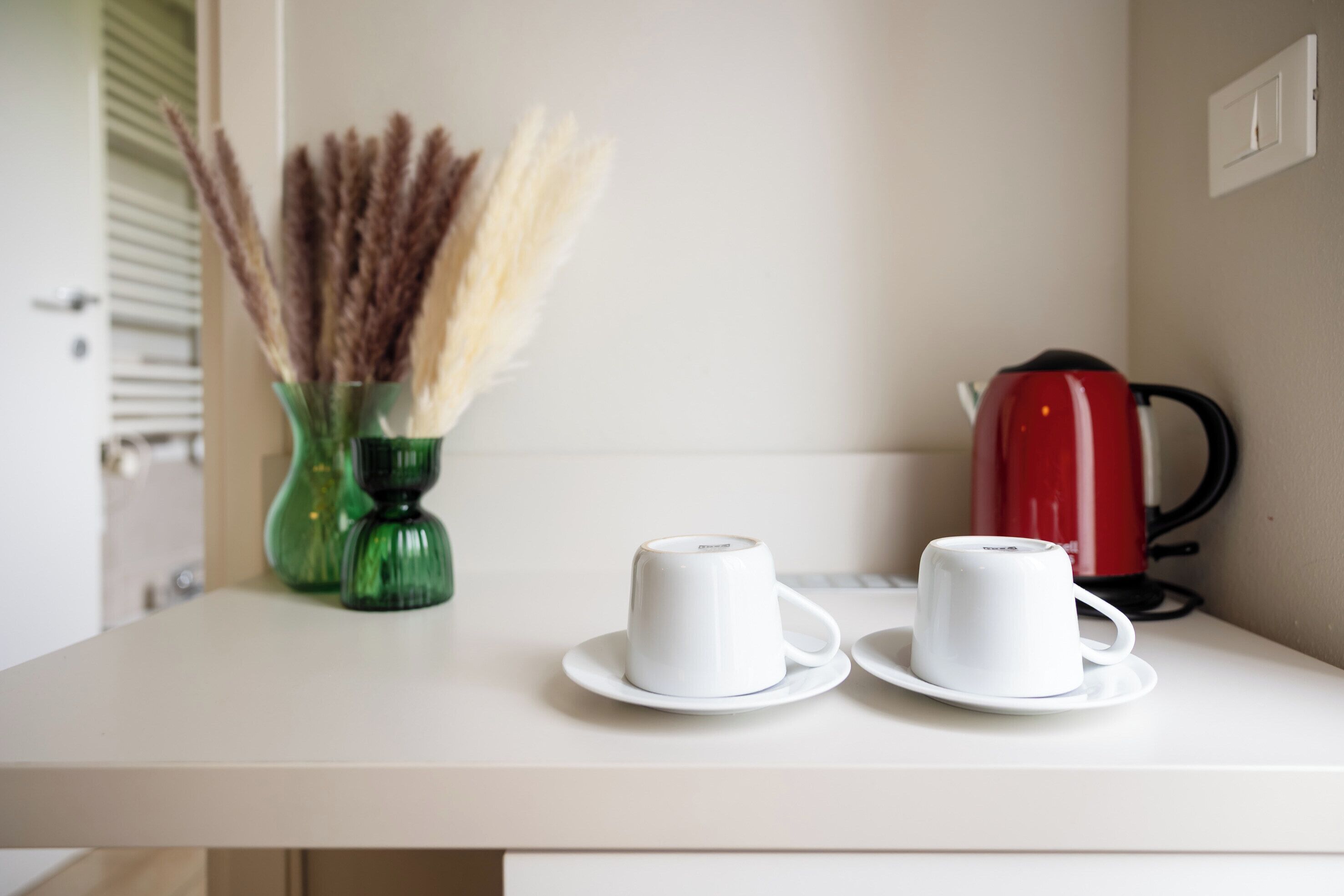 A white table with two white cups on saucers, a green vase with dried plants, and a red kettle.