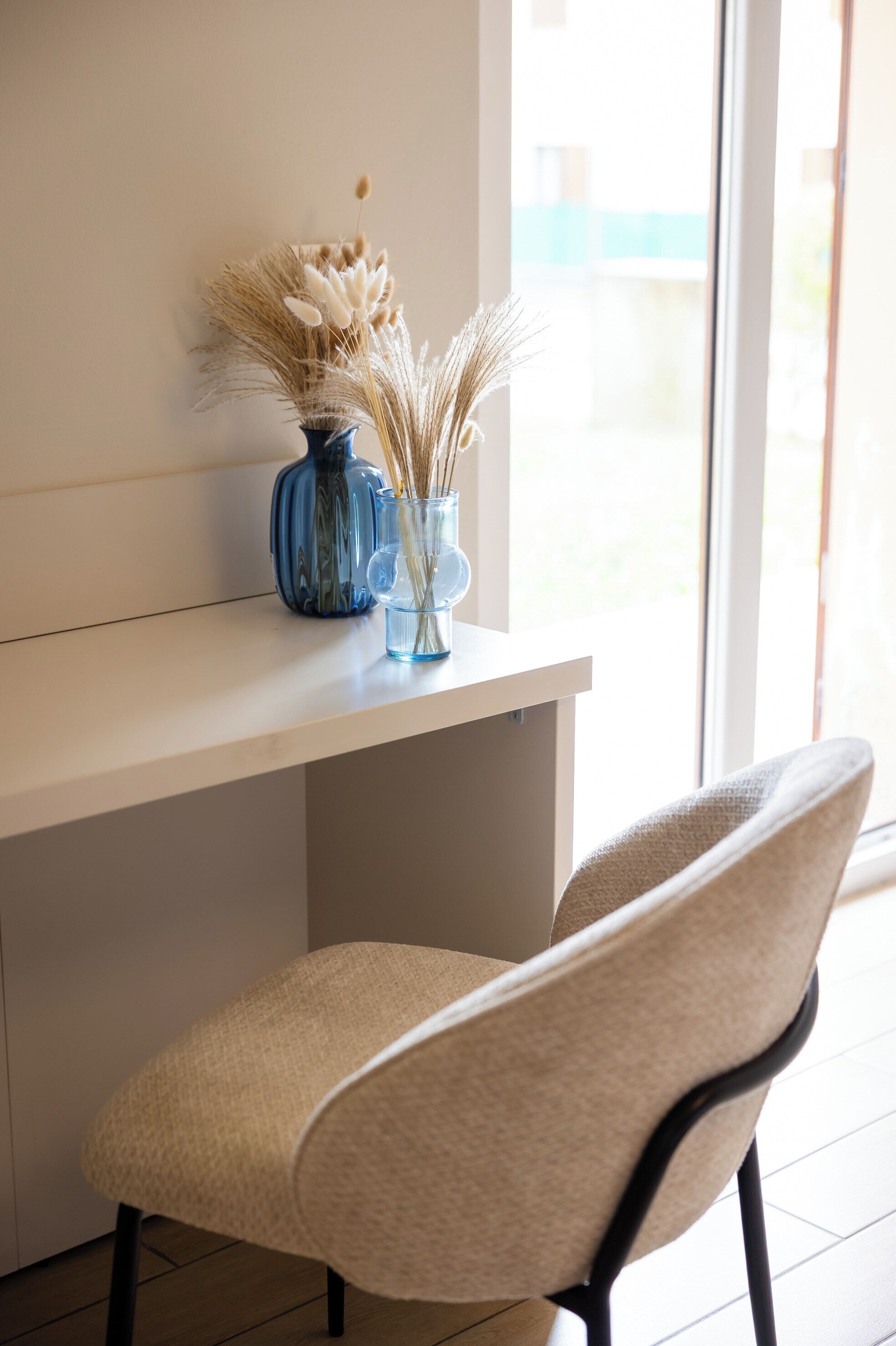A chair and a vase with dried plants on a white table near a window.