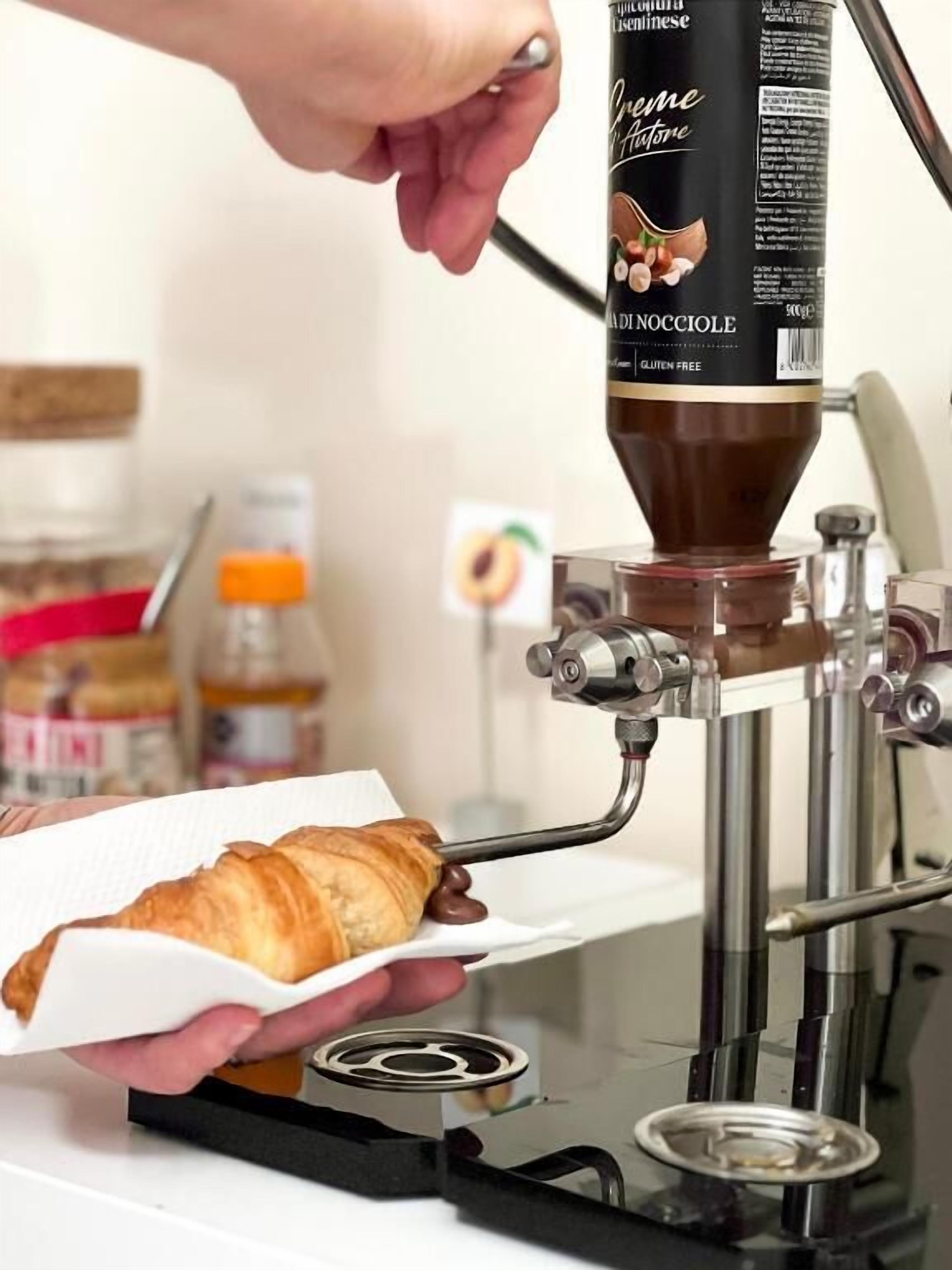 A hand holding a croissant under a coffee machine dispensing a chocolate filling.