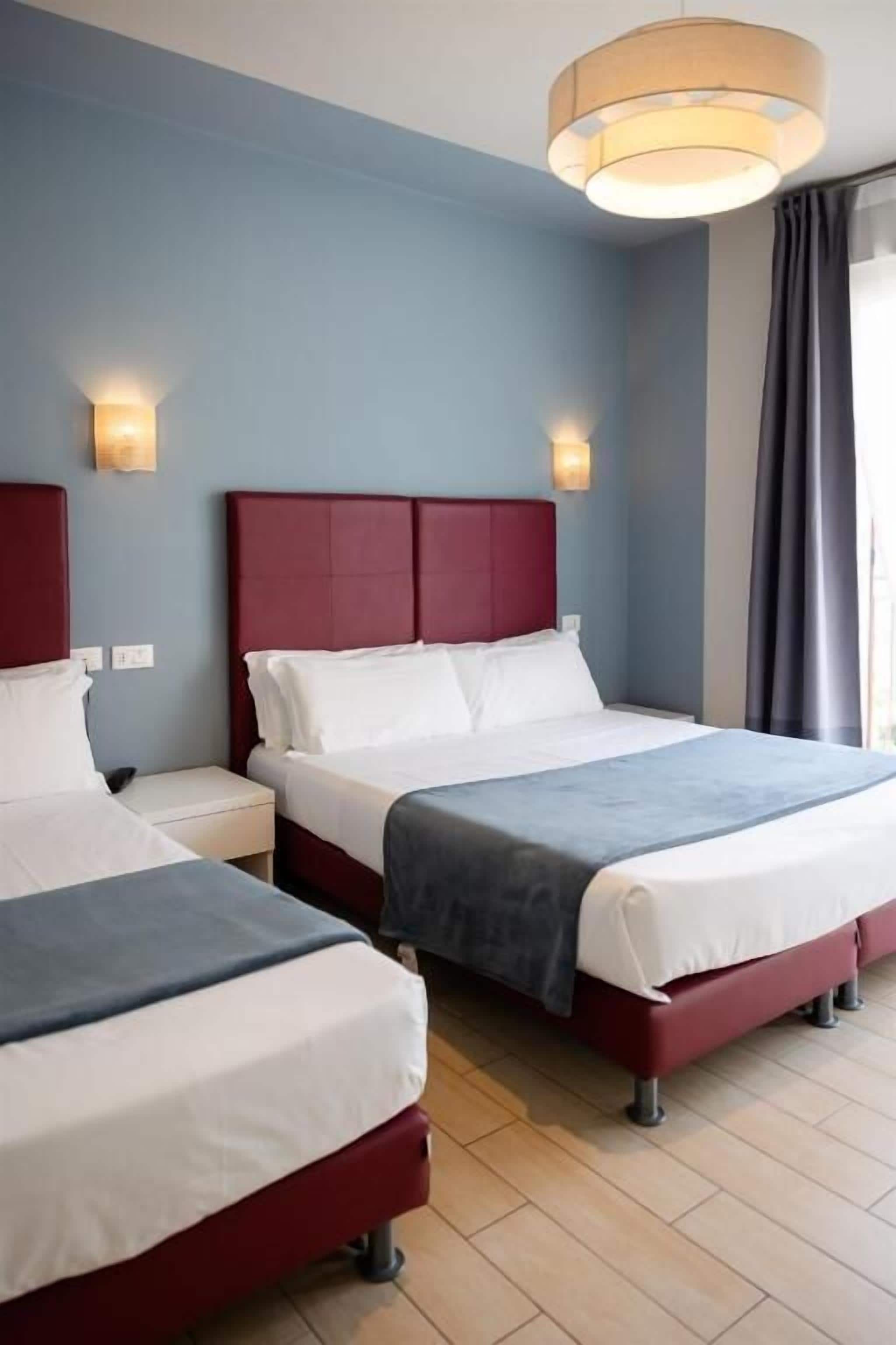 A hotel room with two beds, a red headboard, white bedding, a wooden floor, and a large window with curtains.