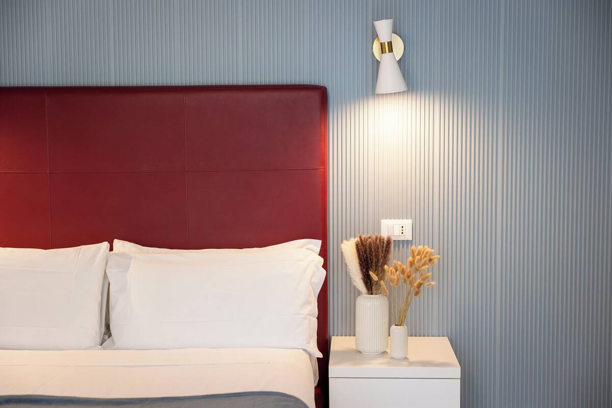 Una cama con cabecera roja, ropa de cama blanca, una mesita de noche blanca con un jarrón de flores secas y una lámpara de pared.