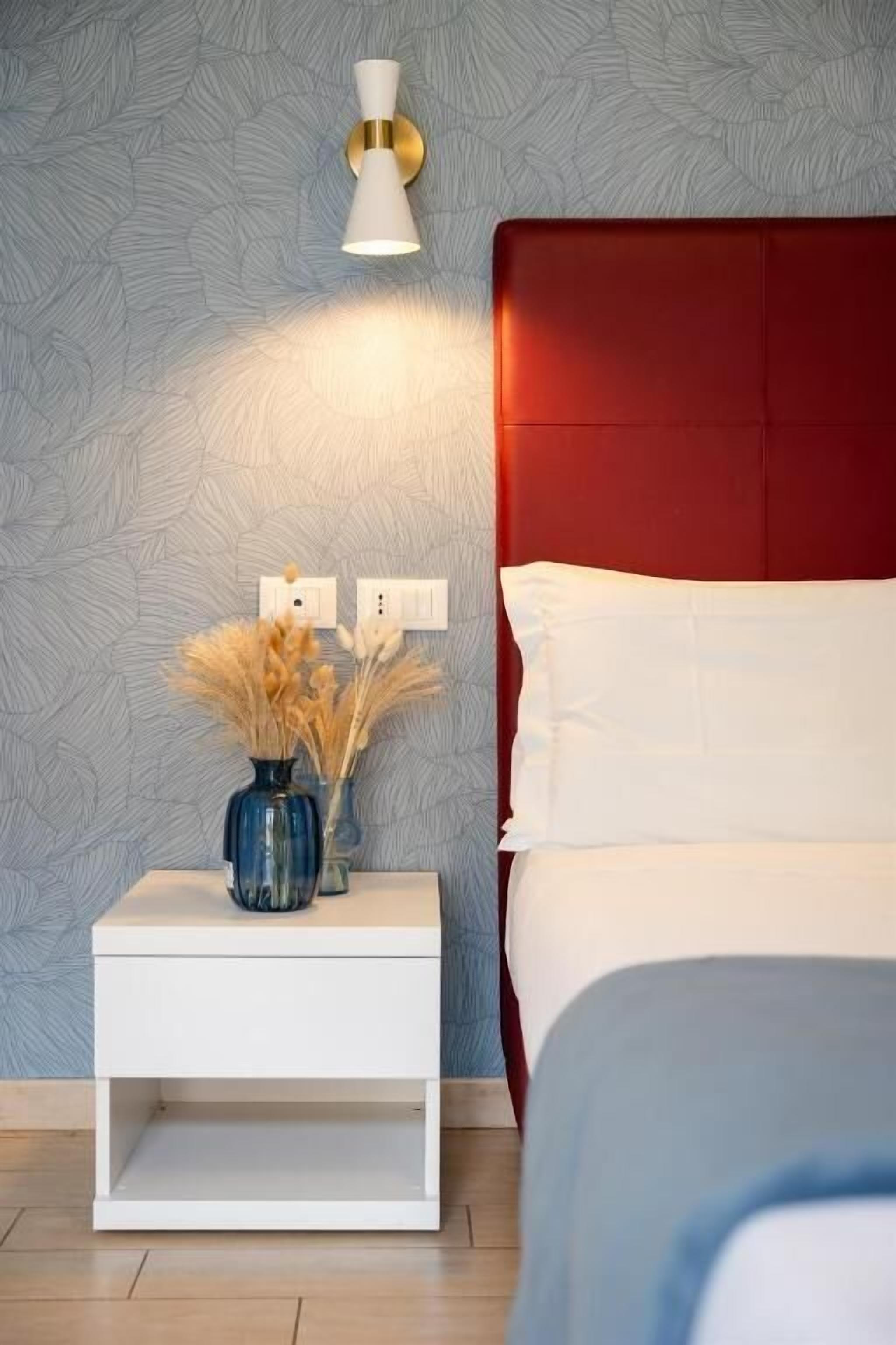 A bedroom with a red headboard, white bedding, a white nightstand with a vase of dried flowers, and a wall-mounted light fixture.