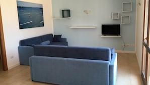 A modern living room with a blue sofa, a flat-screen TV mounted on the wall, and a painting of a beach scene. - Herons Bay Resort (Marsala)