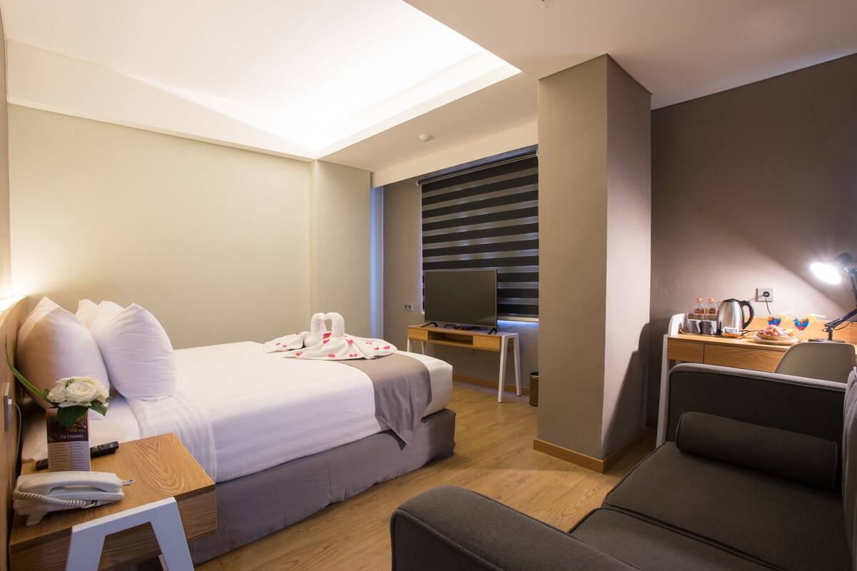 A hotel room with a bed, bedside table, TV, desk, chair, and a small table with a vase of flowers.