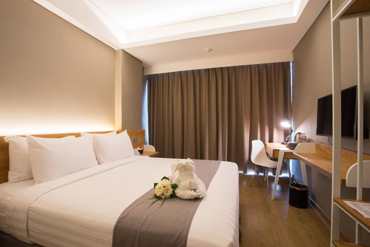 A hotel room with a neatly made bed, a desk with a chair, a television, and a window with curtains.
