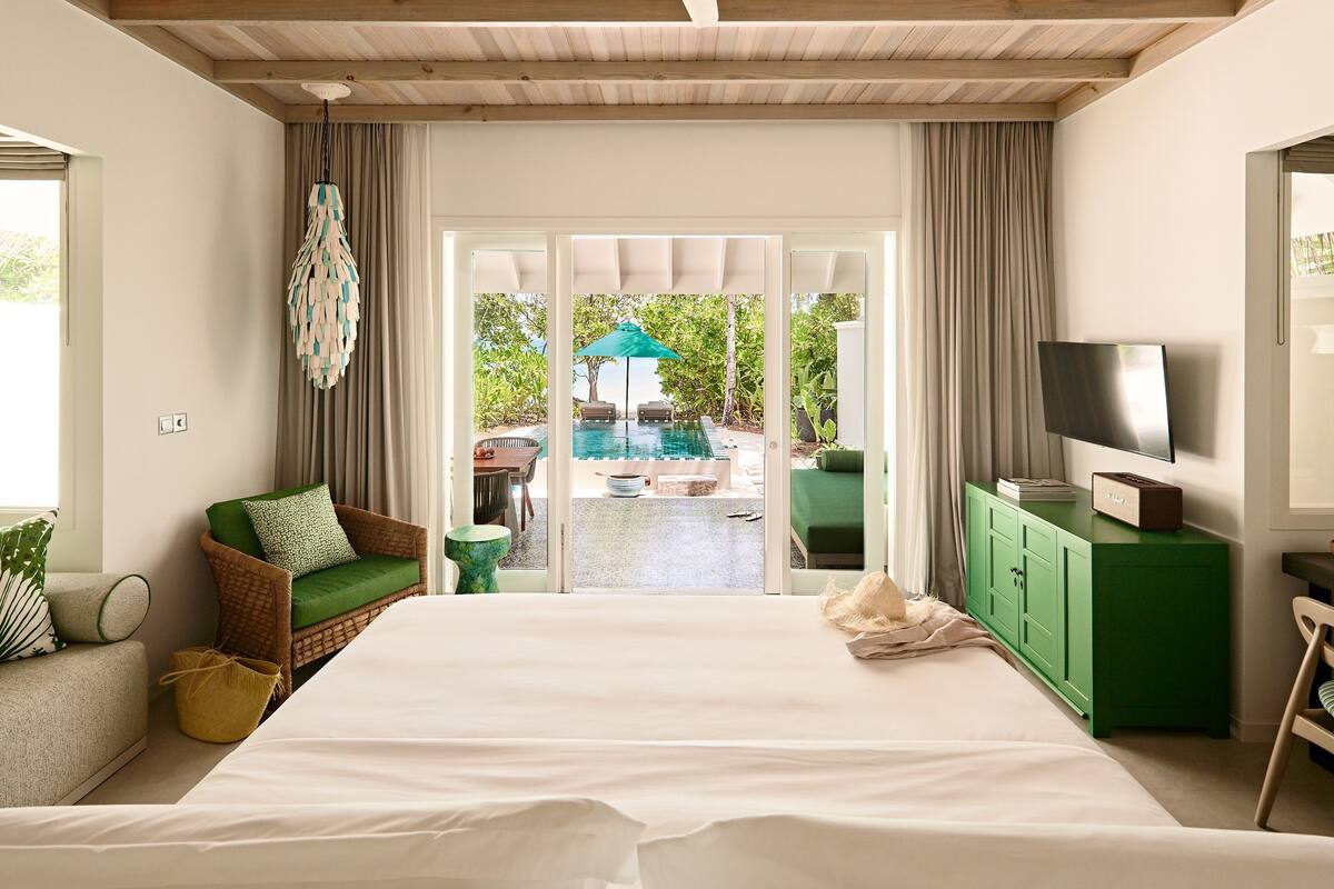Une chambre d’hôtel avec un grand lit, un canapé vert, une télévision et une vue sur l’espace extérieur abritant une piscine.