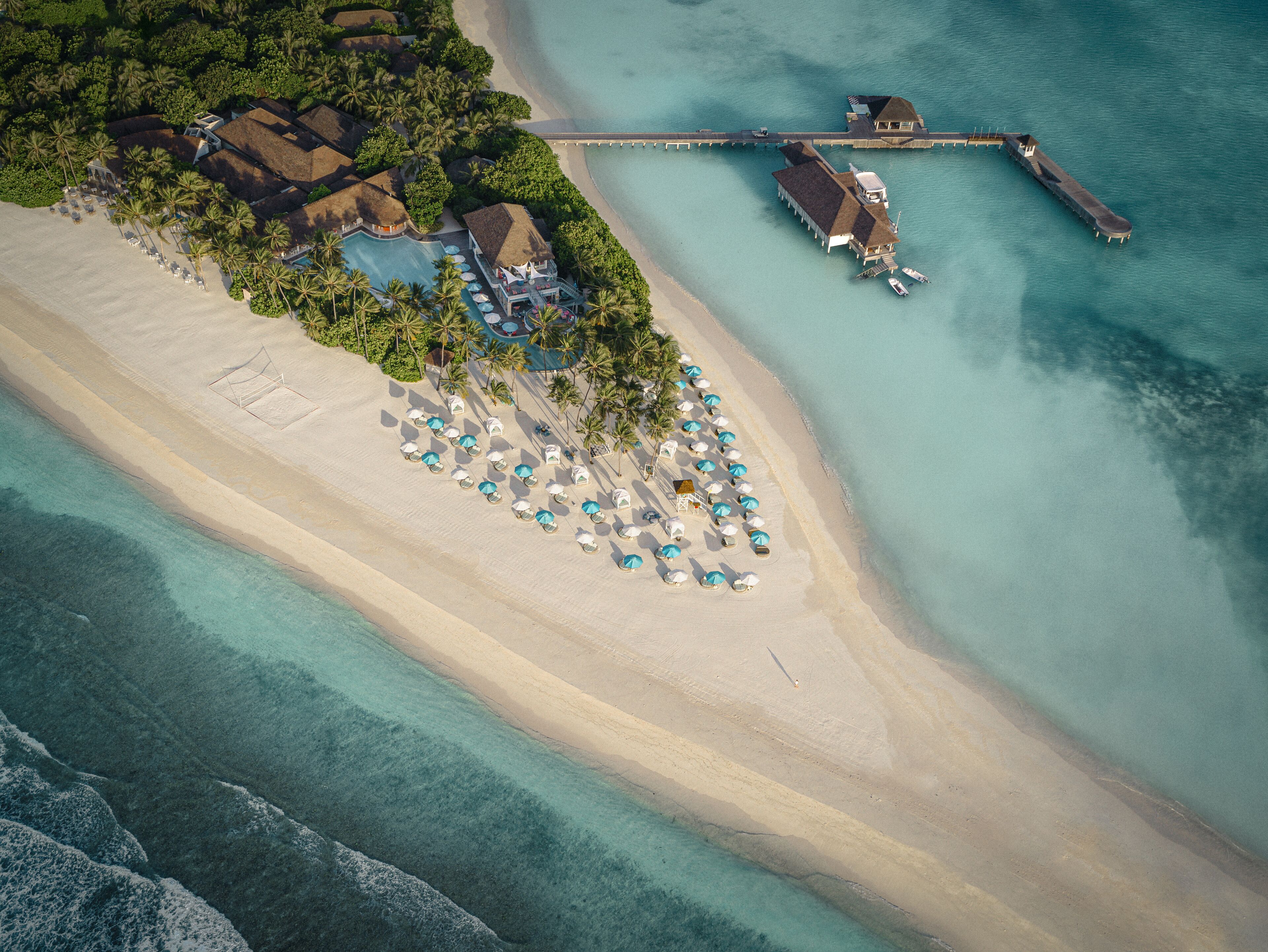 Vista aérea de um resort à beira-mar com um píer extenso, várias bangalôs sobre a água e uma praia arenosa bem cuidada, com guarda-sóis.