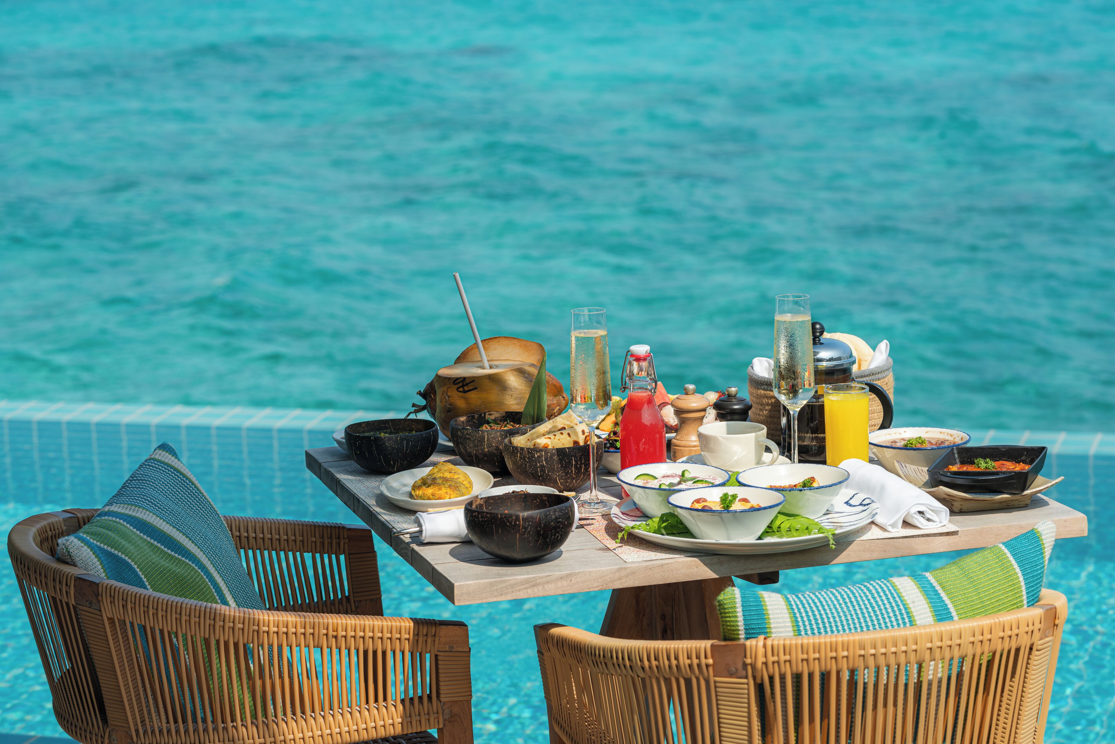 Mesa de comedor con diversos platos y bebidas junto a una piscina, con sillas de mimbre y vistas al agua.
