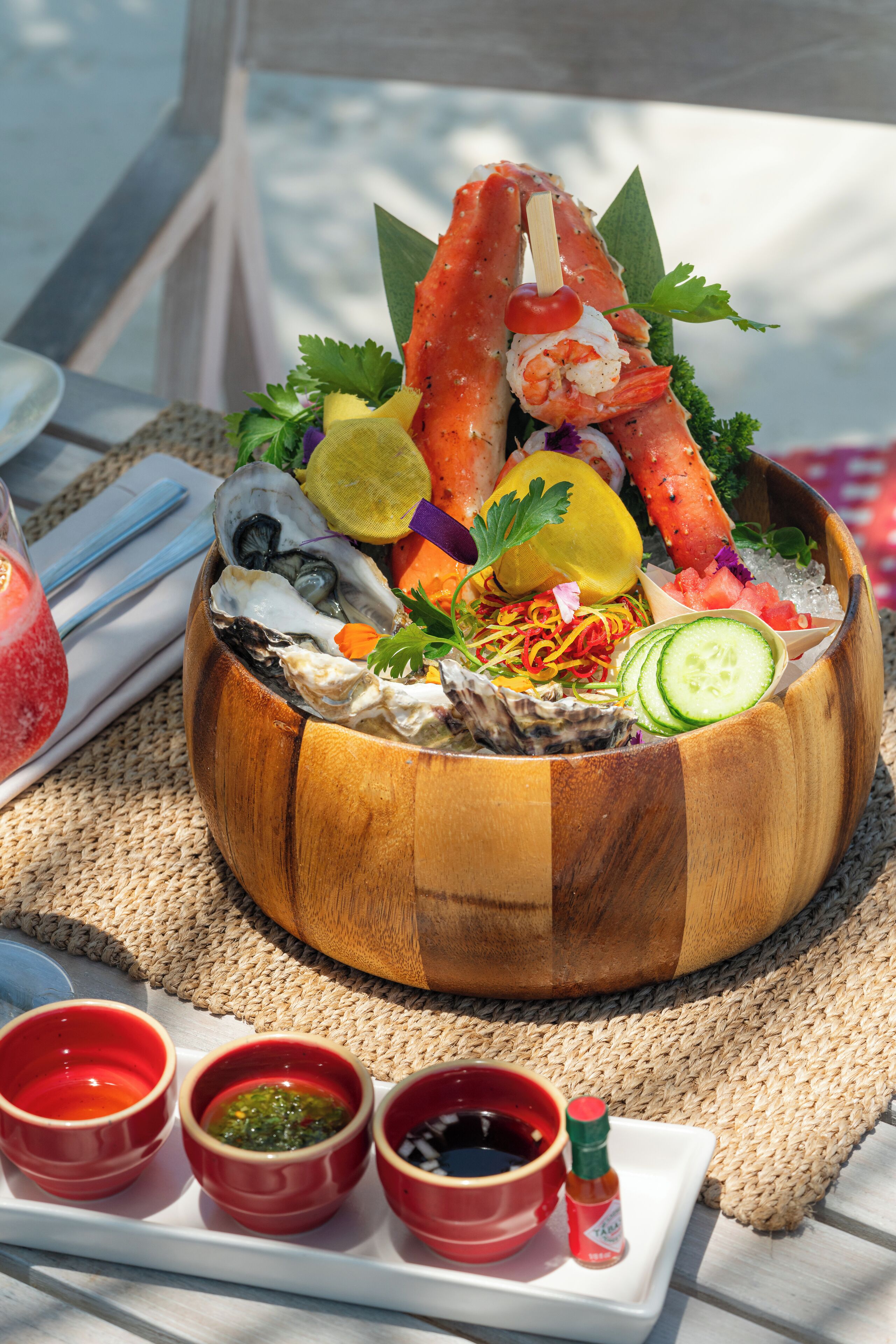 Un tazón de madera lleno de mariscos, como camarones y ostras, adornado con vegetales y hierbas, acompañado de tres recipientes pequeños rojos con salsas sobre un tapete tejido.