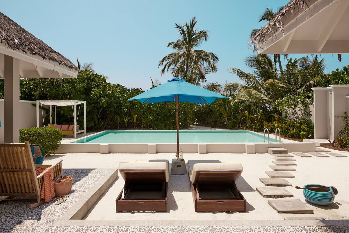 Un espace aménagé au bord de la piscine, avec des chaises longues, un parasol bleu et une vue dégagée sur une piscine entourée de palmiers.