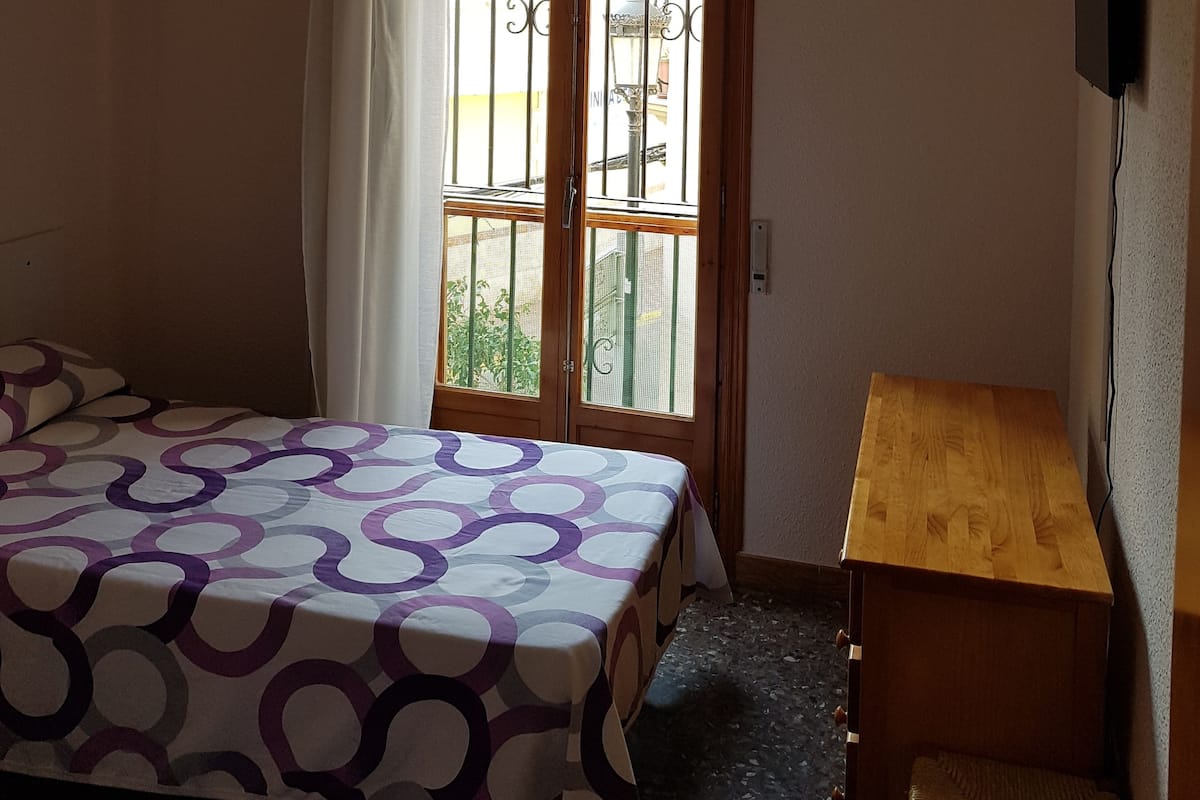Un dormitorio con una cama, una mesita de noche de madera, una ventana con vistas y aire acondicionado instalado en el techo.