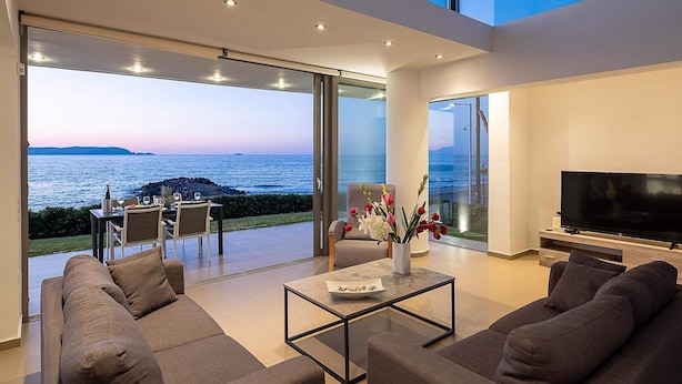 Una sala de estar moderna con un televisor grande, asientos cómodos y un ventanal que da a un balcón con vistas al mar.