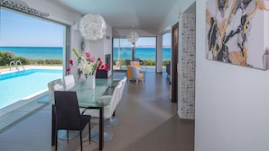 Un comedor moderno con mesa y sillas de vidrio, vista al mar y un cuadro abstracto grande en la pared.