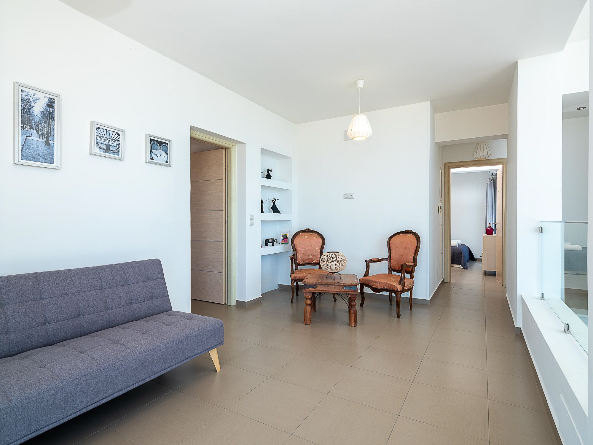 Ein modernes Wohnzimmer mit einer grauen Couch, zwei Holzsesseln, einem kleinen Tisch und einer Hängeleuchte. Regale mit Dekoration und eine Tür zu einem anderen Raum sind ebenfalls zu sehen.