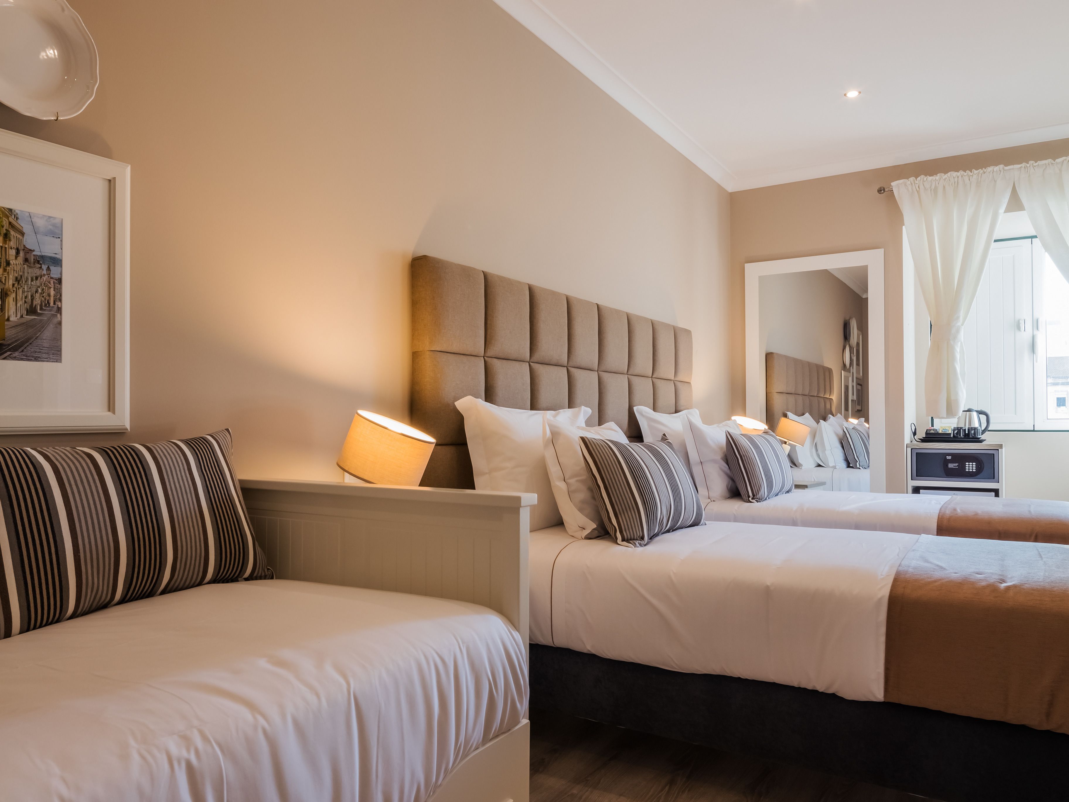 A hotel room with two beds, a bedside table with a lamp, a framed picture on the wall, and a window with curtains.