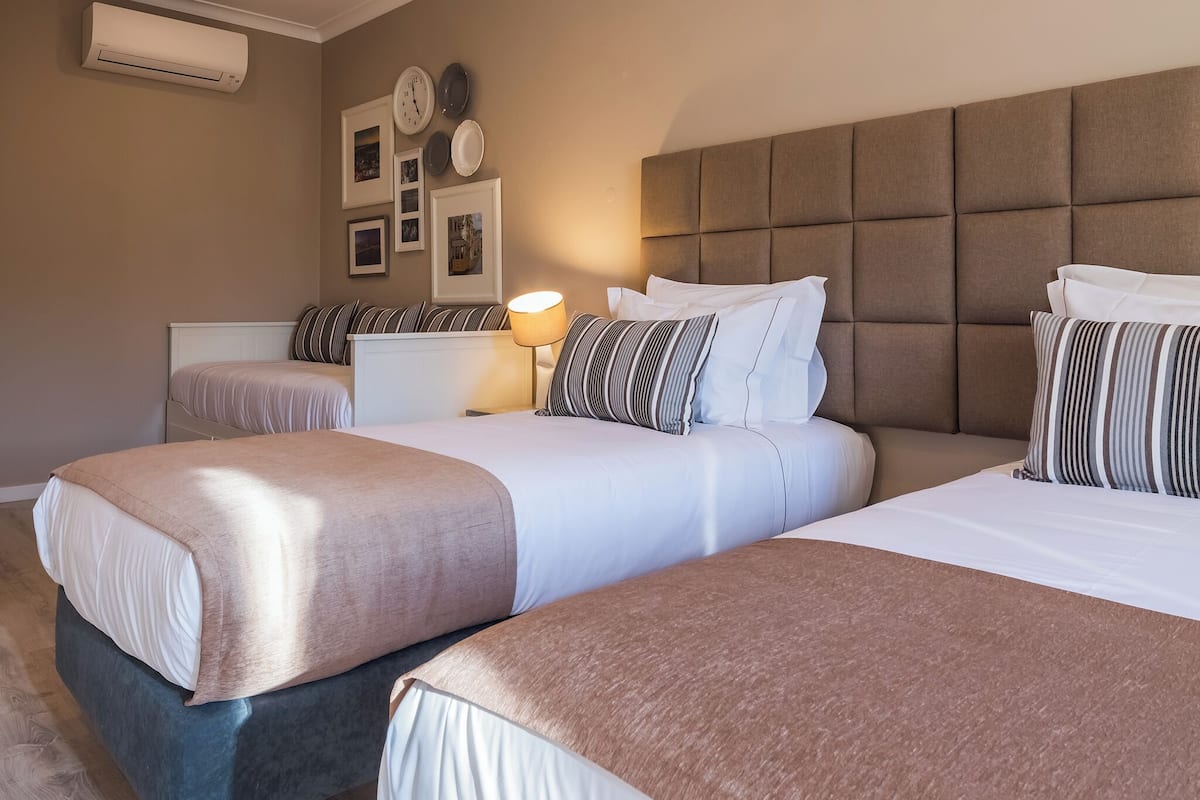 A hotel room with two beds, a bedside table with a lamp, a wall-mounted air conditioner, and framed pictures.