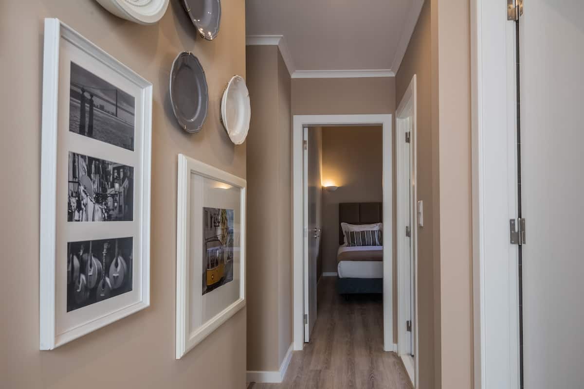 A hallway with framed photographs on the walls, a bed in the adjacent room, and a door at the end.