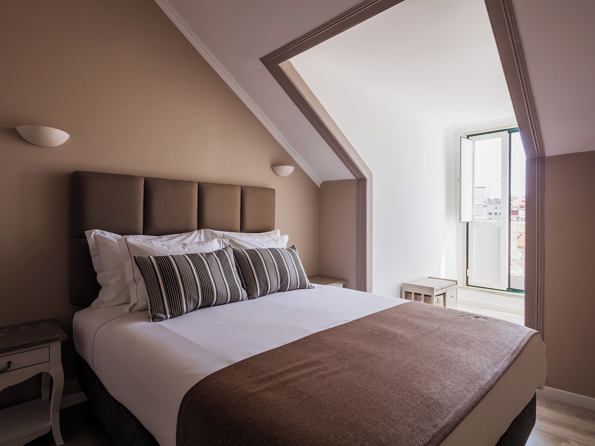 Ein Schlafzimmer mit einem großen Bett, einem Kopfteil mit drei Feldern, einem Nachttisch und einem Fenster mit Blick auf Gebäude.