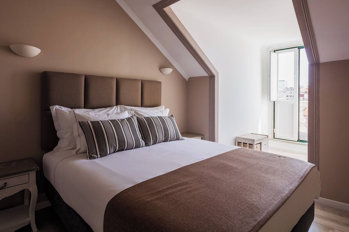 A bedroom with a large bed, a headboard with three panels, a bedside table, and a window with a view of buildings.