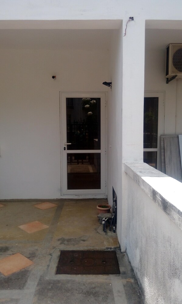 A small, enclosed outdoor area with a white door leading to a dark interior. There is a concrete floor with a metal grate, a potted plant, and a metal bench.