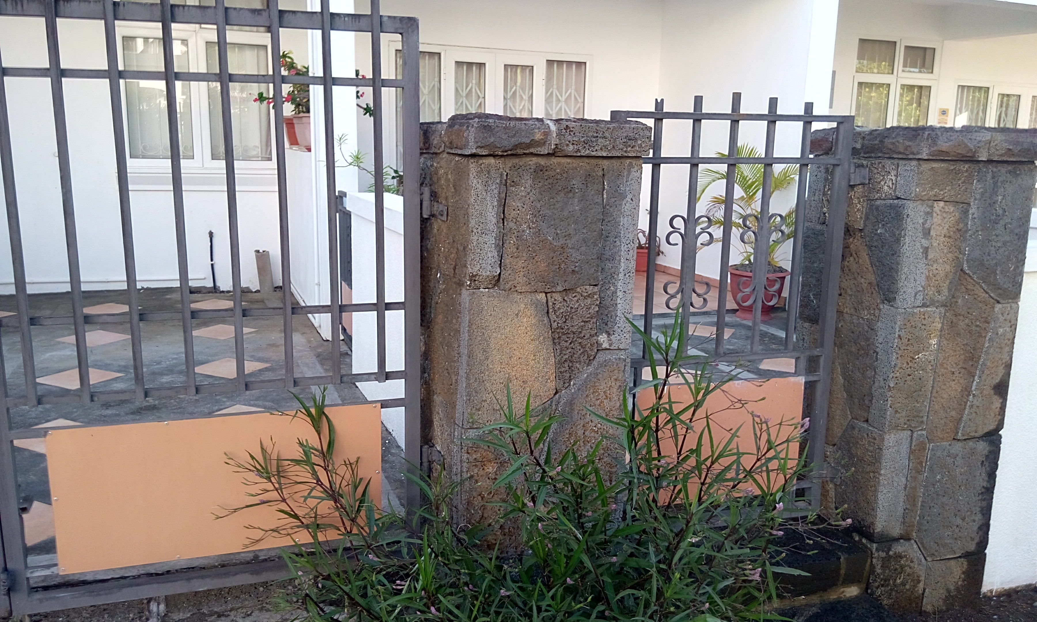 Uma entrada protegida por portões de pedra e grades de metal, que dá acesso a um pátio com piso de azulejos e plantas em vasos.