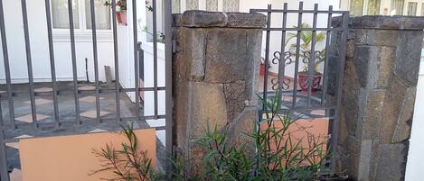 Uma entrada protegida por portões de pedra e grades de metal, que dá acesso a um pátio com piso de azulejos e plantas em vasos.