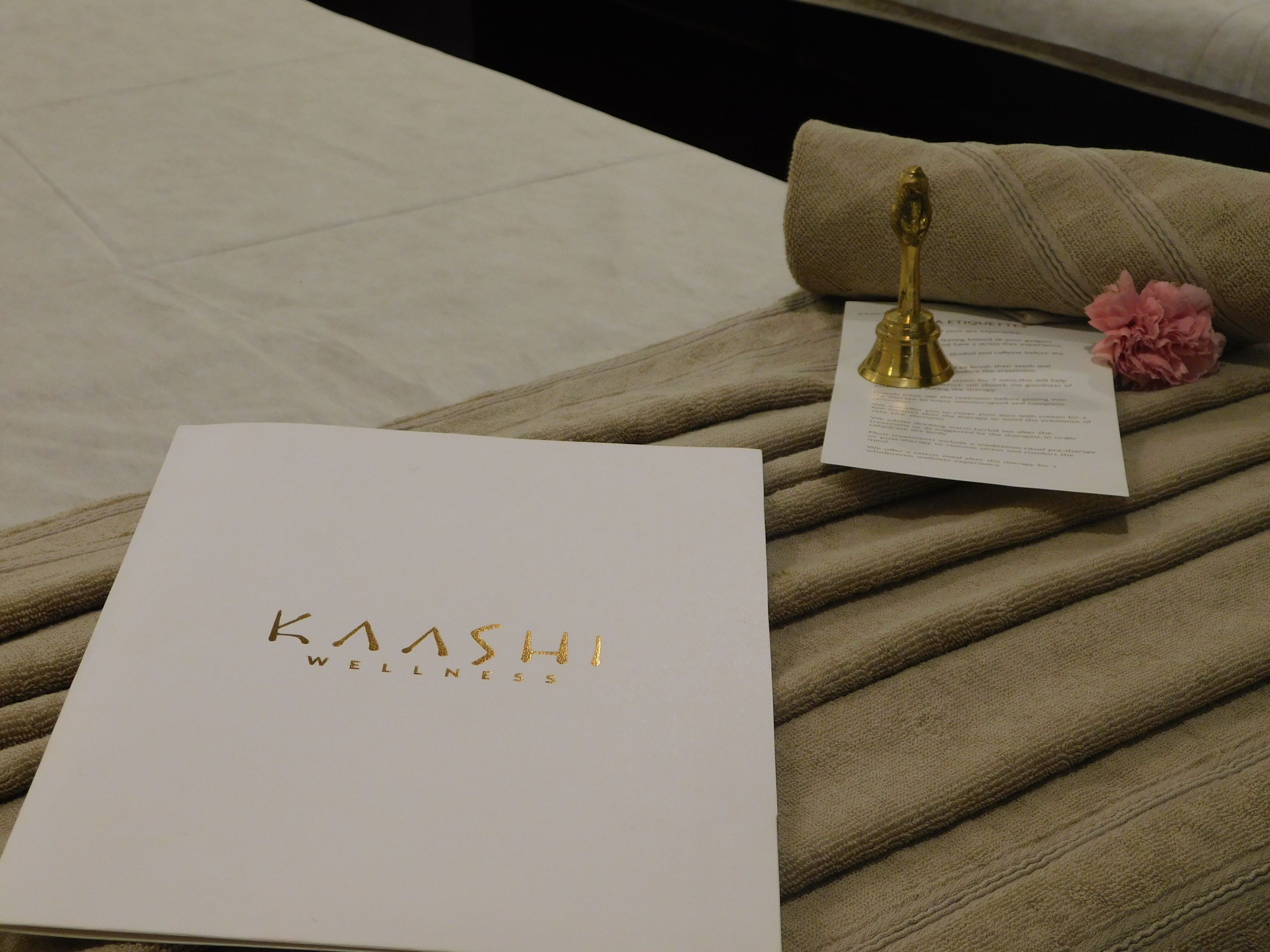 Un petit livret blanc avec le texte « KAASH WELLNESS », une cloche dorée, une fleur rose et un morceau de papier sur une surface beige texturée.