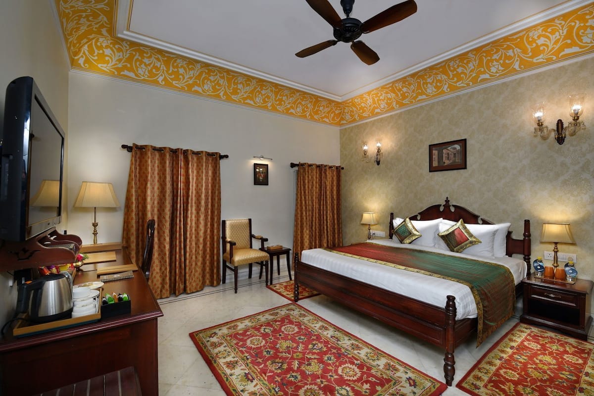 A hotel room with a bed, a desk with a coffee maker, a chair, a television, and a ceiling fan.