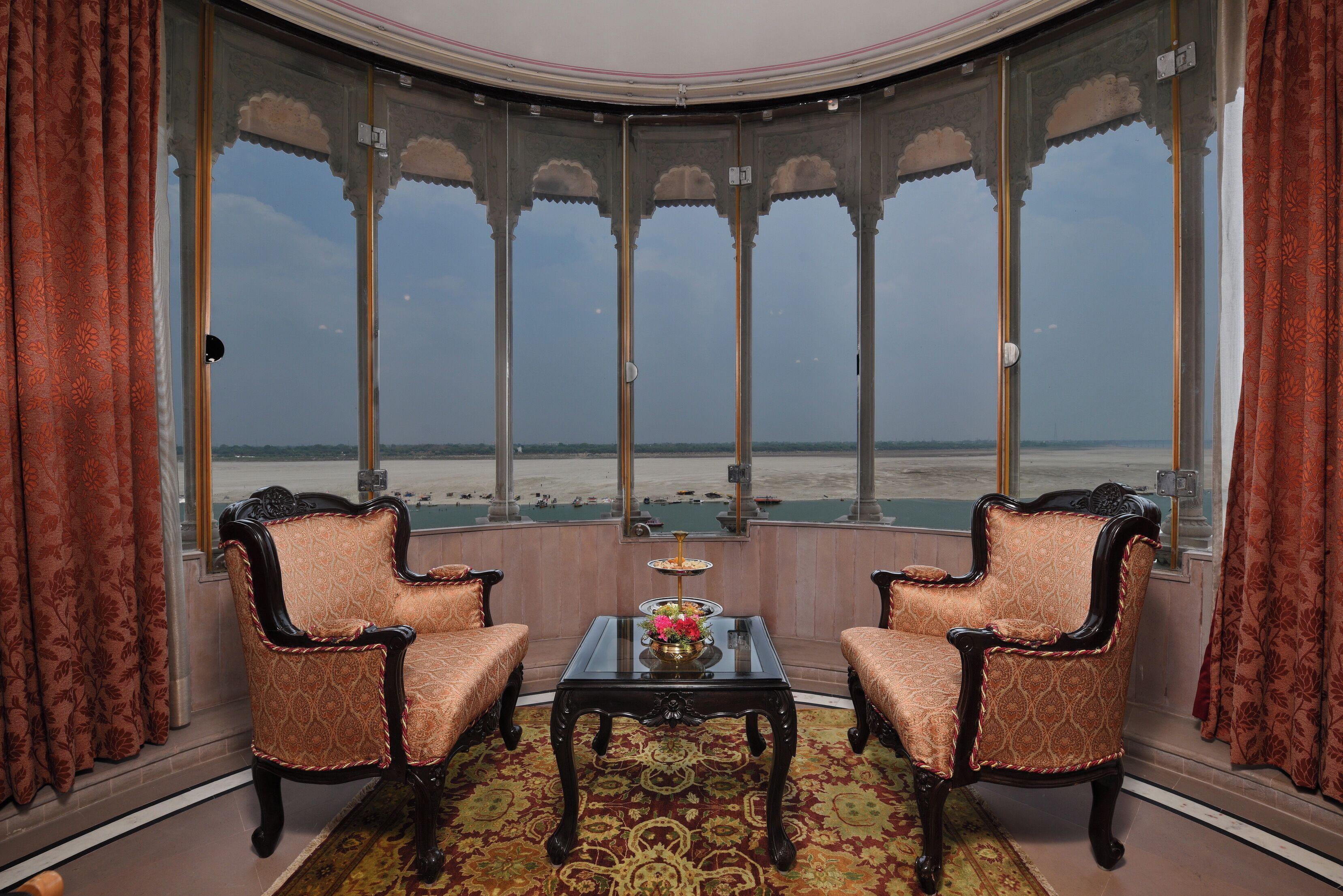 Une pièce dotée de deux fauteuils richement décorés, d’une table à plateau en verre et offrant une vue sur la plage grâce à une grande fenêtre en arc de cercle.