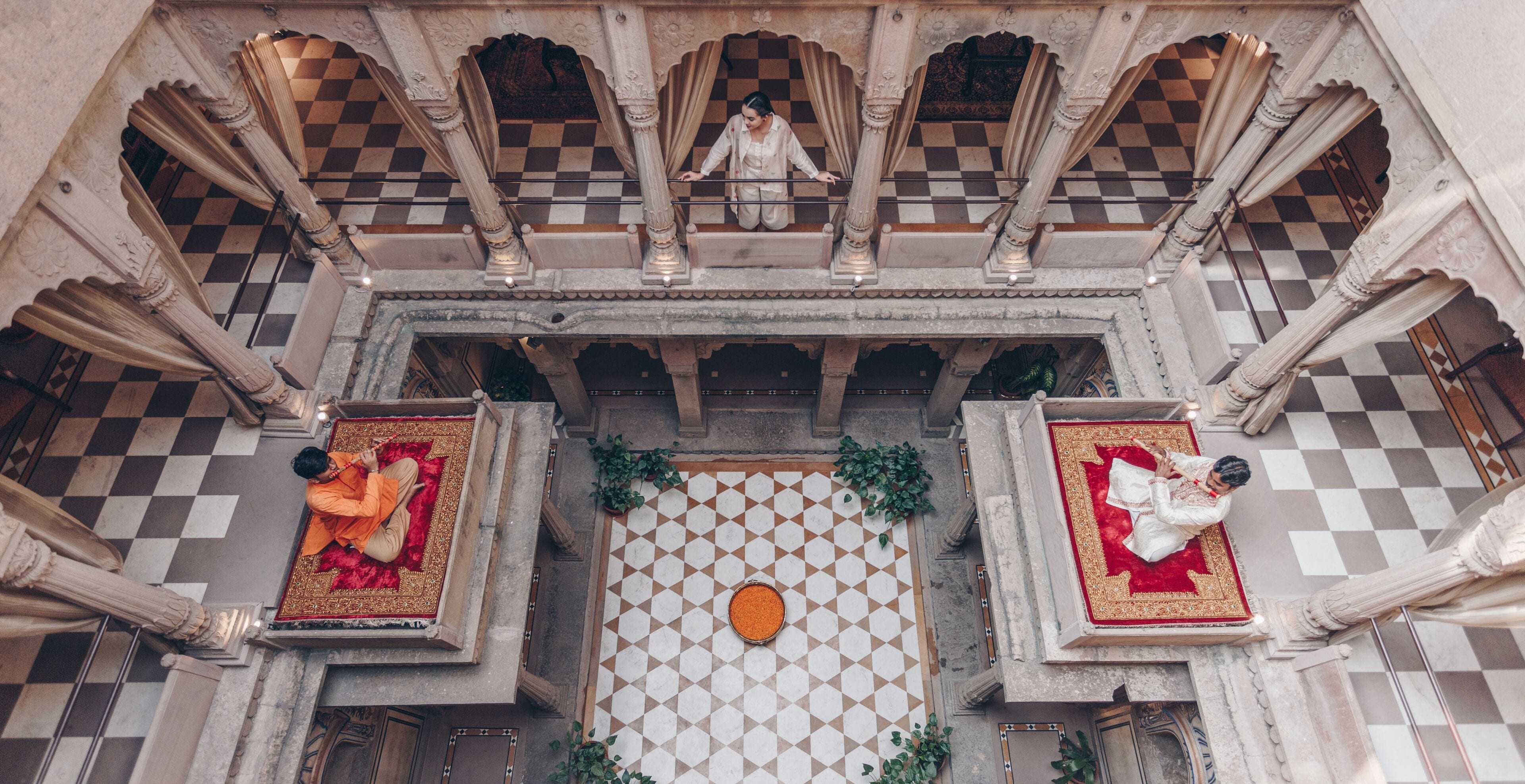 A courtyard with a checkered floor, ornate columns, and two individuals seated on red carpets.