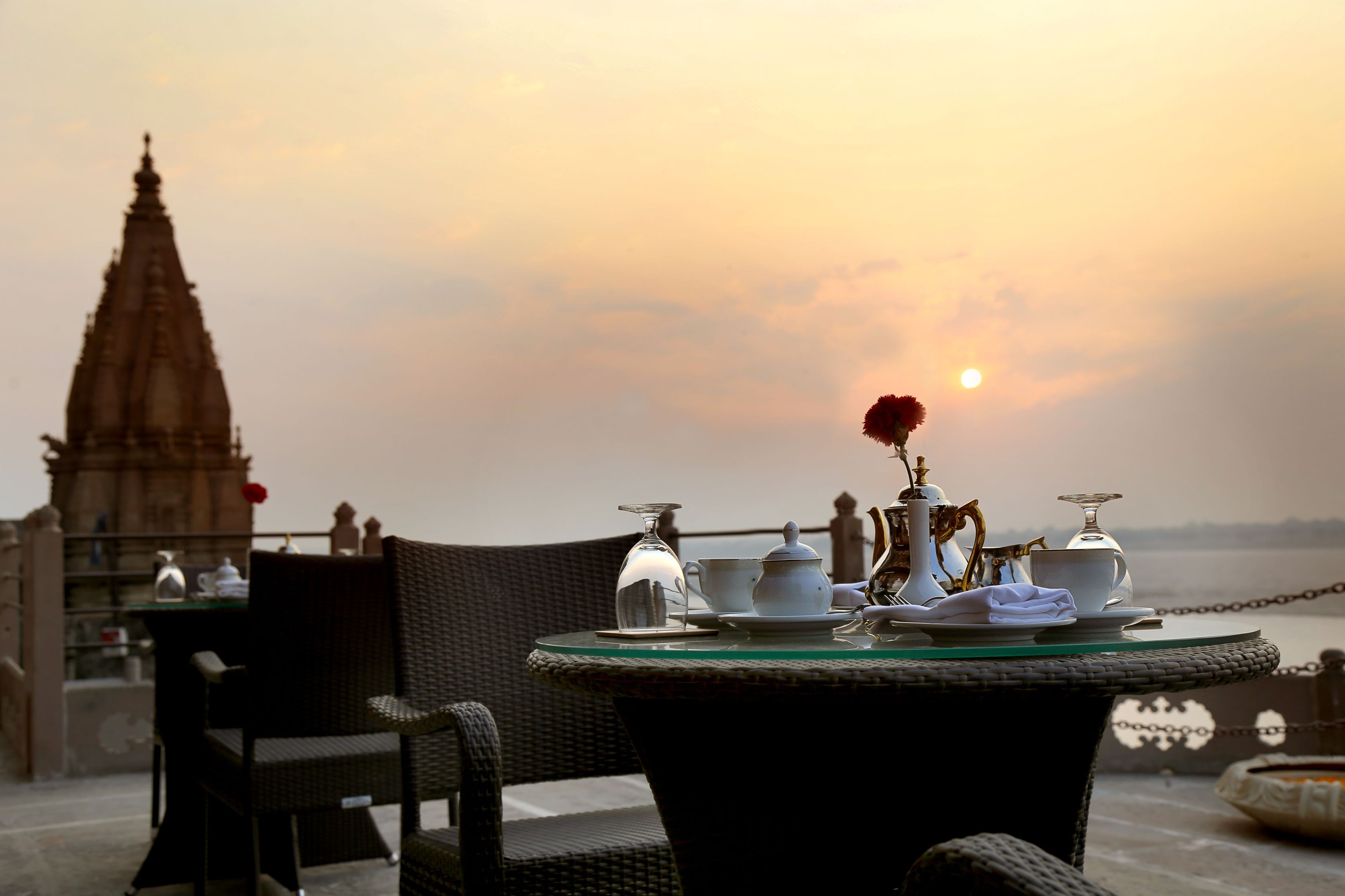 A table set with tea service and a vase with a flower, overlooking a body of water and a temple in the background.