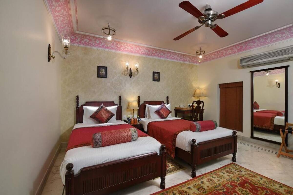 A bedroom with two wooden beds, a ceiling fan, wall sconces, a mirror, and a red carpet.