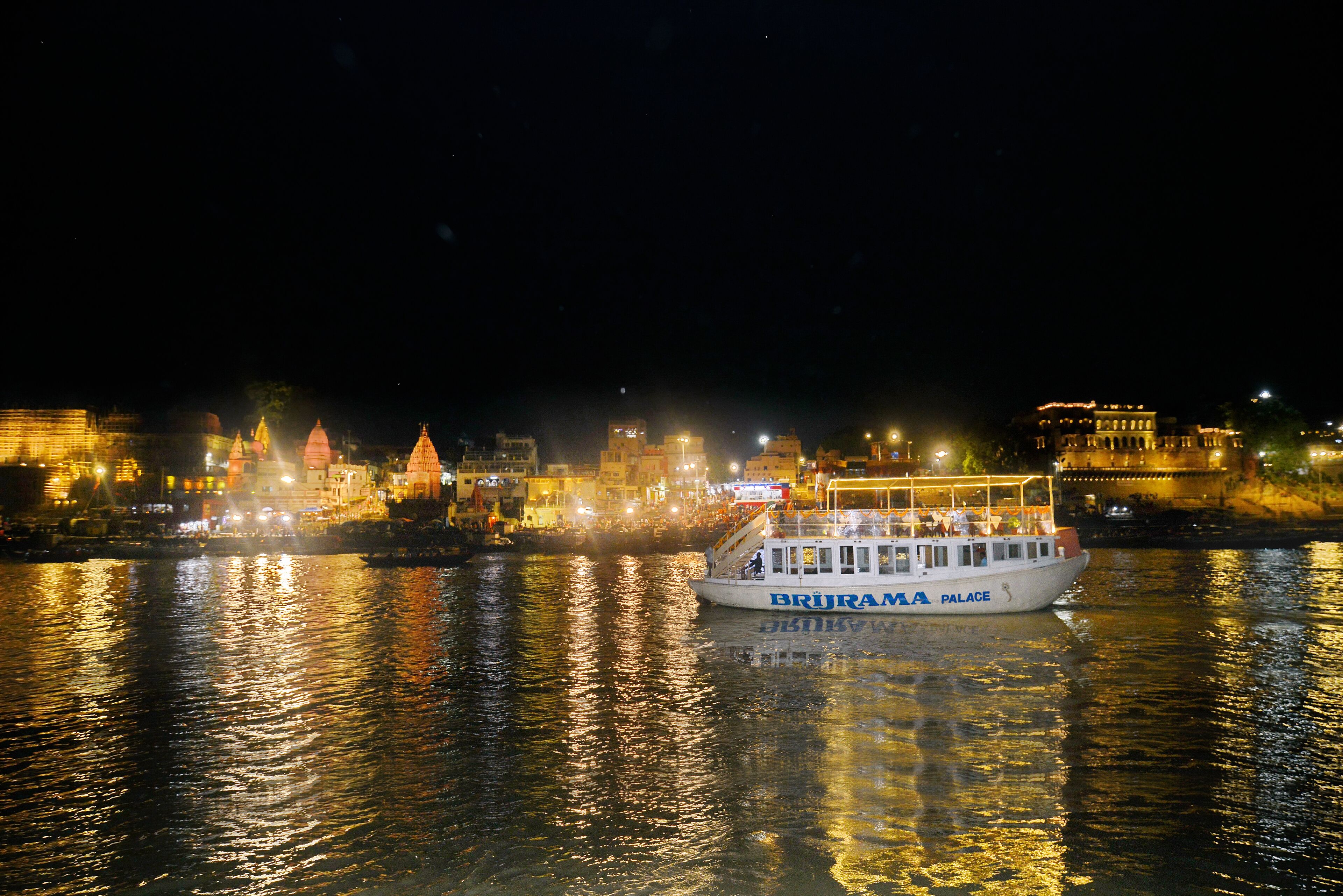 Un barco llamado BRIJAMPA PALACE en un río de noche, con edificios iluminados al fondo.