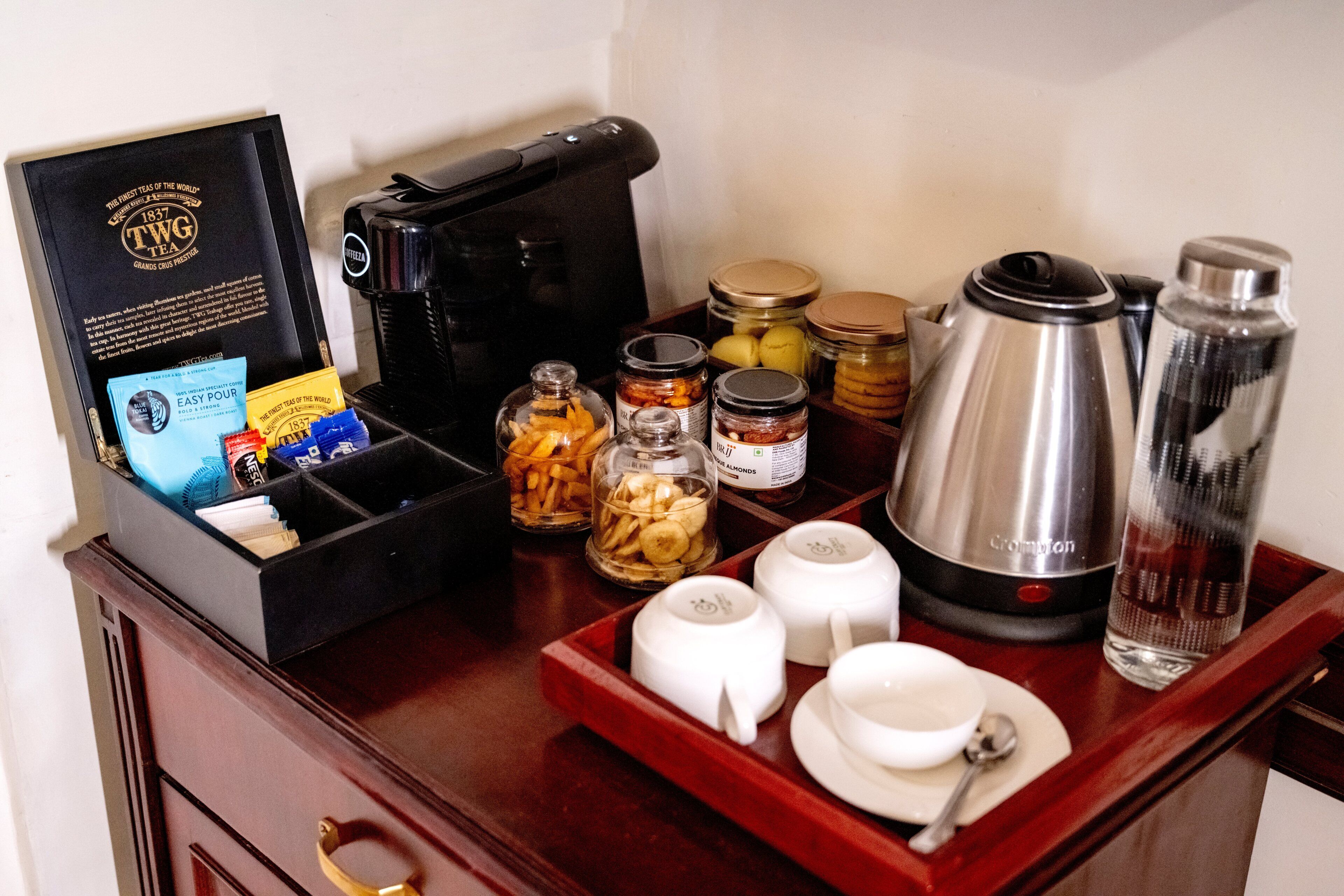 A coffee station with a coffee maker, tea packets, jars of condiments, and a water bottle.