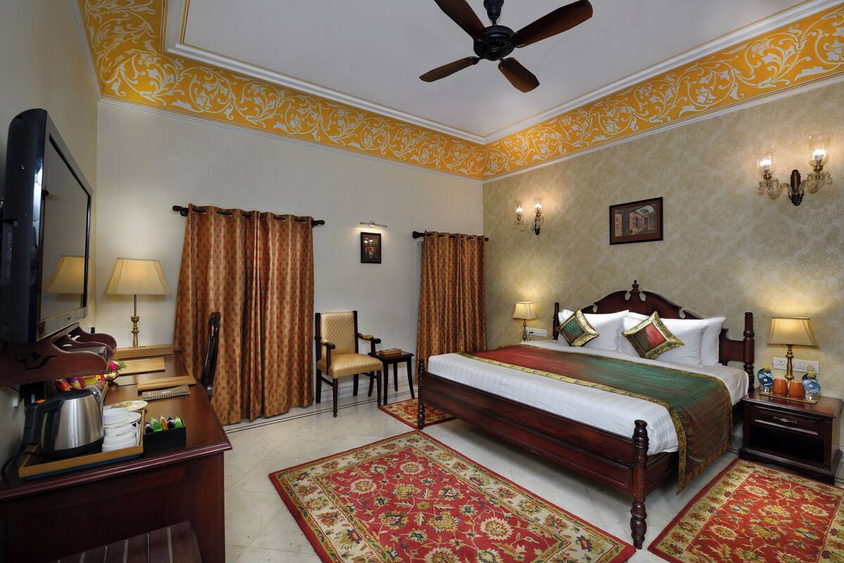 A hotel room with a bed, a desk with a coffee maker, a chair, a nightstand with lamps, and a ceiling fan.