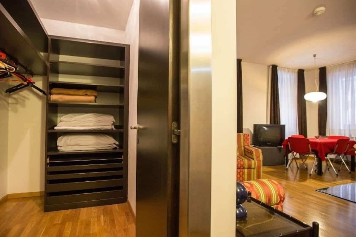 A hotel room with a wooden floor, a dining area with a table and chairs, a television, and a closet with hanging clothes.