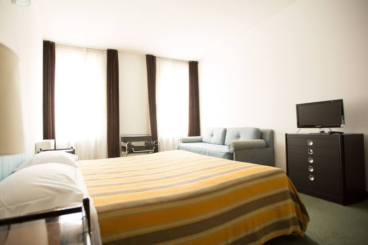 A hotel room with a bed, a sofa, a TV on a cabinet, and a window with curtains.
