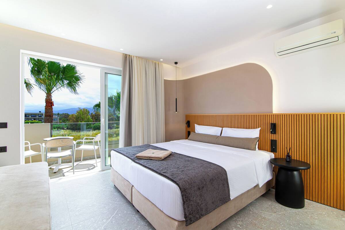 Une chambre d’hôtel moderne avec un grand lit, une petite table, une chaise et un balcon offrant une vue sur les palmiers et le paysage urbain.