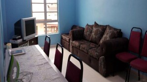 A room with a blue wall, a sofa with patterned cushions, a television on a stand, and a table with chairs.