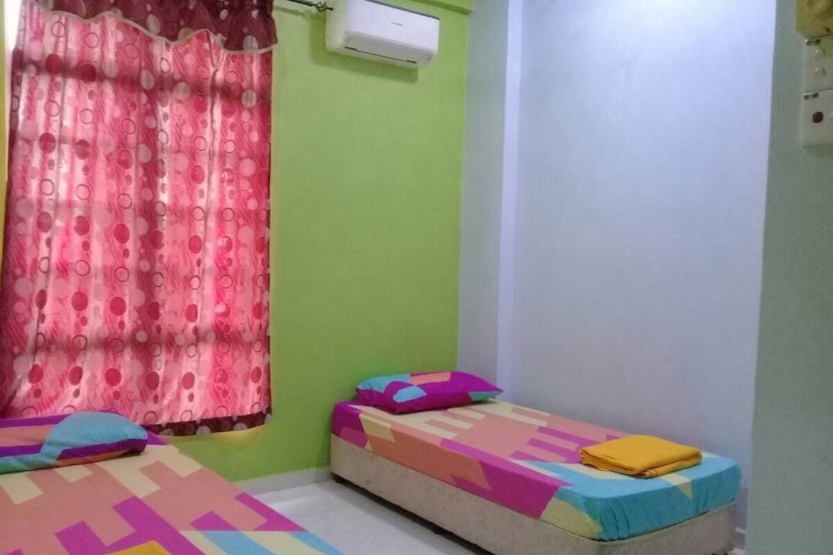 Une petite pièce aux couleurs vives, avec deux lits, chacun recouvert d’une couverture colorée. Il y a une fenêtre avec un rideau rose et un appareil de climatisation fixé au mur.