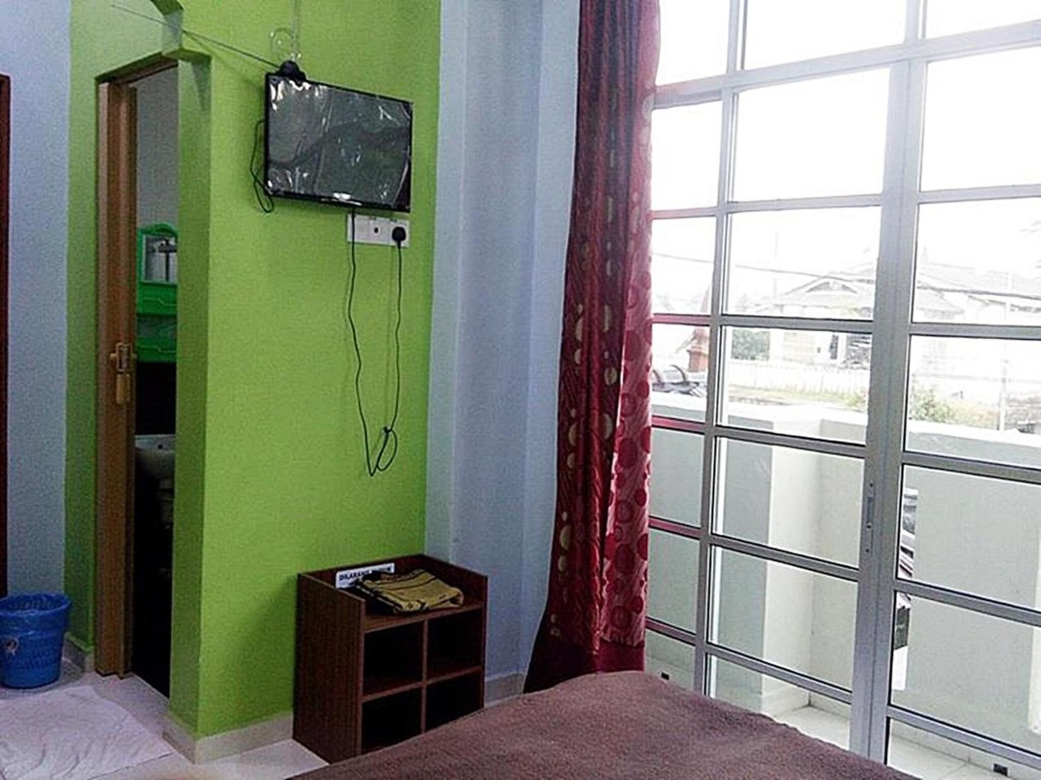 Une pièce comprenant un lit, un téléviseur fixé au mur, une fenêtre avec des rideaux et une petite étagère.