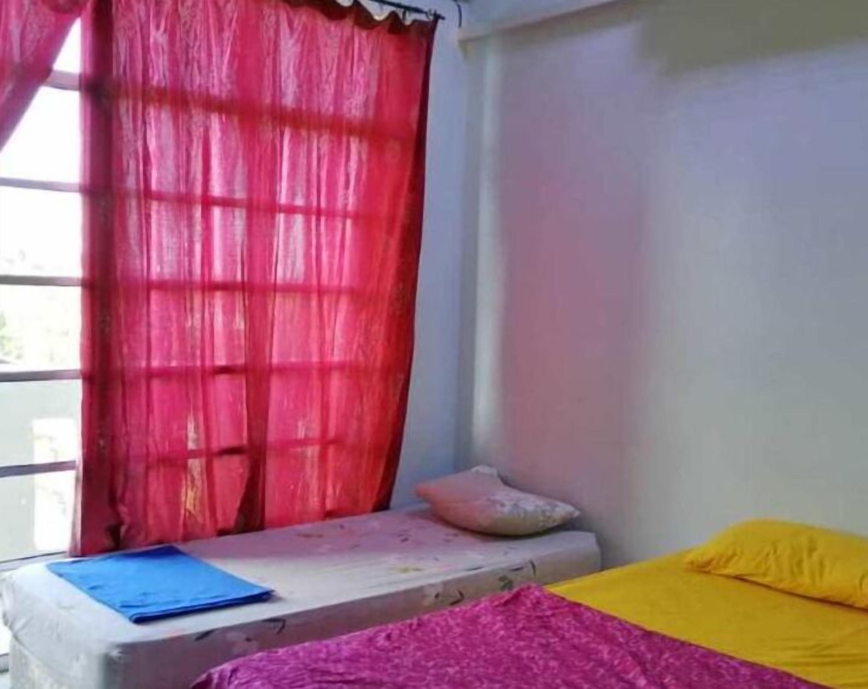 Ein Zimmer mit einem Bett, einer kleinen Bank, einem Fenster mit roten Vorhängen und Blick nach draußen.