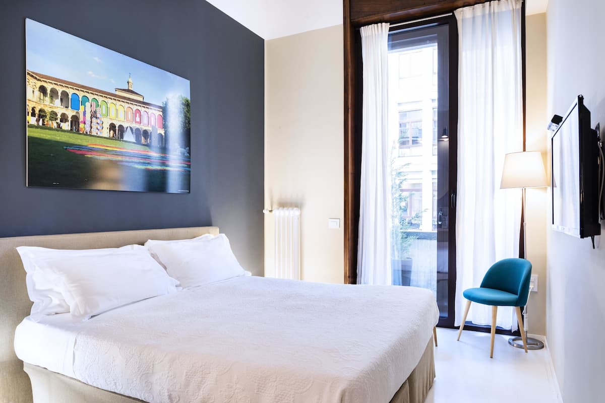 A hotel room with a large bed, a painting of a building, a chair, and a window with curtains.