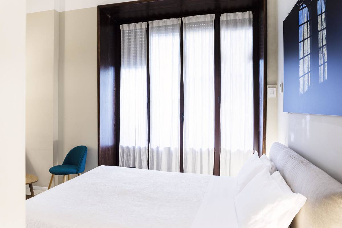 A hotel room with a bed, a chair, and a large window with curtains.