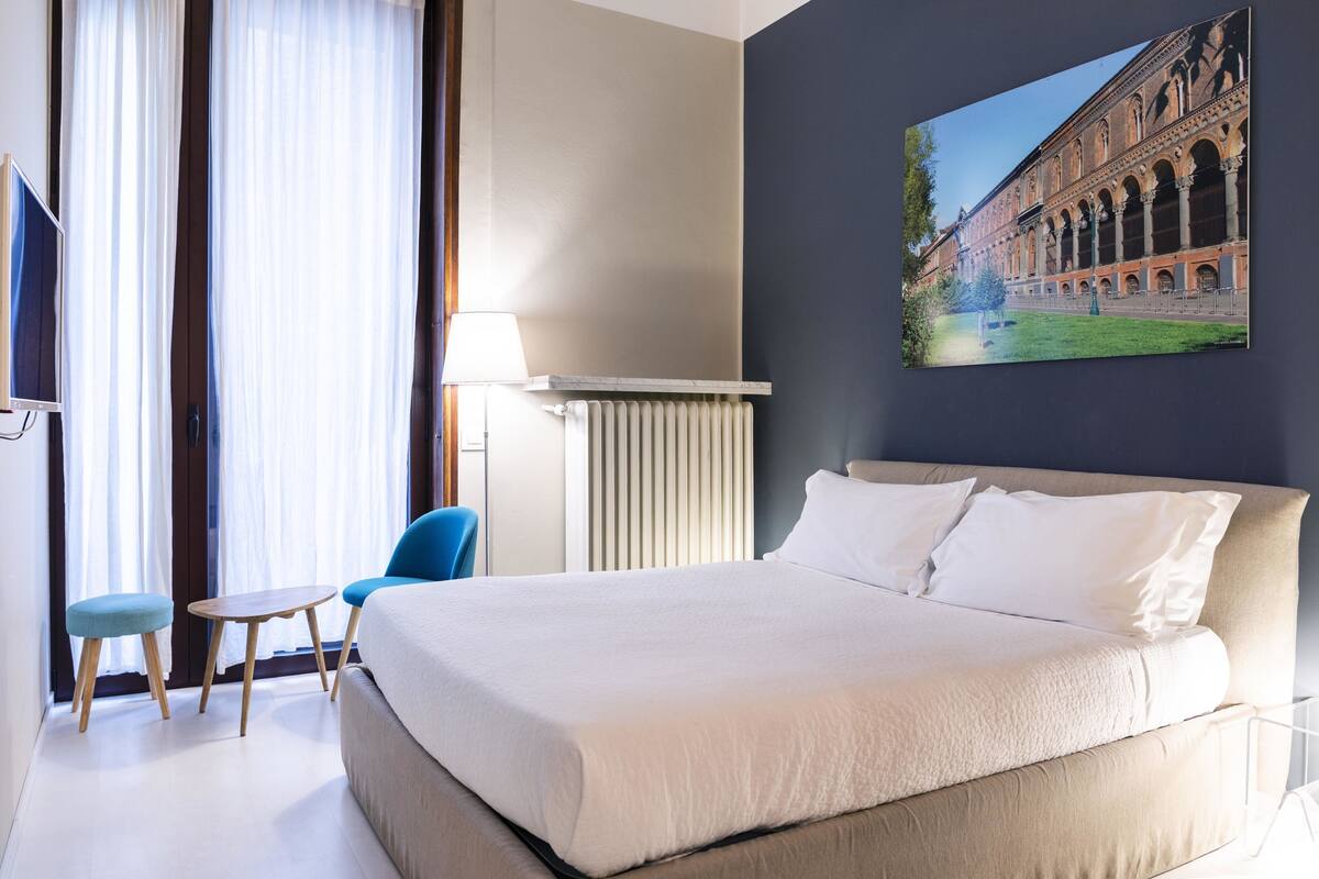 A hotel room with a large bed, a desk, two chairs, a radiator, and a painting of a building on the wall.
