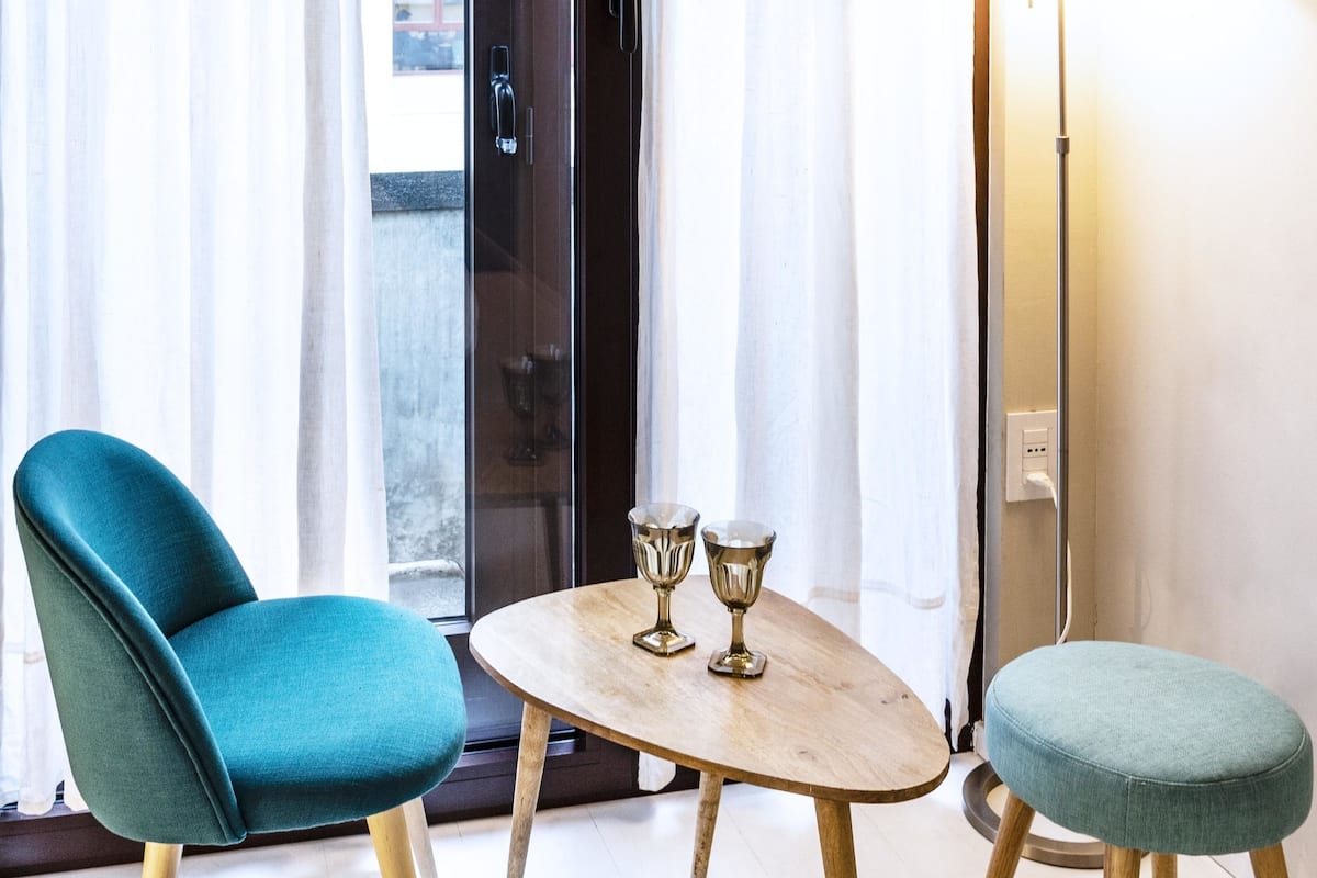 A hotel room with a teal armchair, a small round table with two glasses, and a lamp.