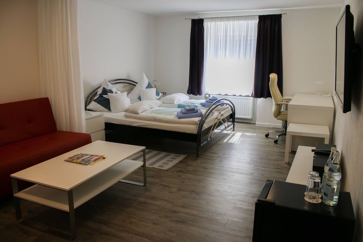 A hotel room with a bed, a red sofa, a white desk, a chair, and a TV.